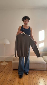 Szary sweter z dzianiny o okrągłym dekolcie, trzymany na wieszaku przez osobę w granatowej koszulce na ramiączkach, w zestawieniu z szerokimi niebieskimi dżinsami.