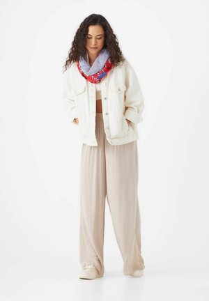 Witte denimjack, beige wijde broek, rode en gemusterde sjaal, witte schoenen. Krullend haar, handen in de zakken, neutrale achtergrond.