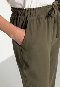 Pantalon taille ajustable vert olive en tissu doux et texturé, avec une poche latérale et un détail de ceinture nouée.