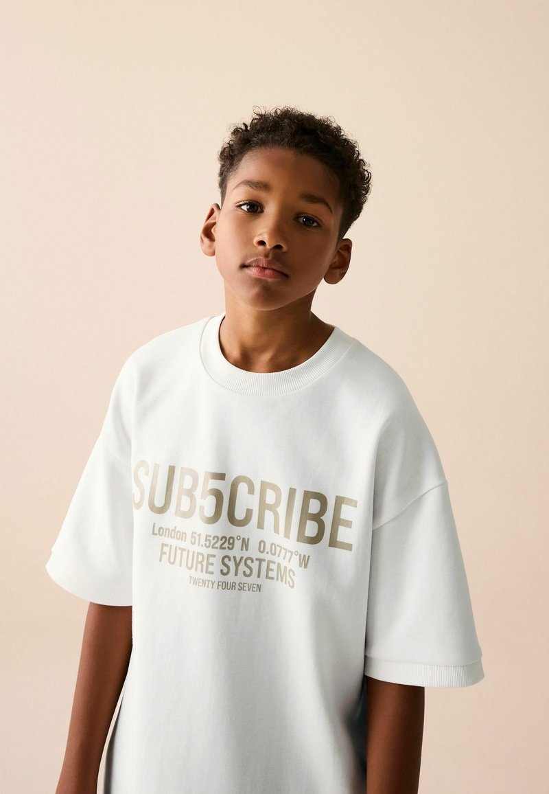 Hvid oversized t-shirt med korte ærmer, der har guldtekst: "SUB5CRIBE," geografiske koordinater, og "FUTURE SYSTEMS." Glat stoffets tekstur.