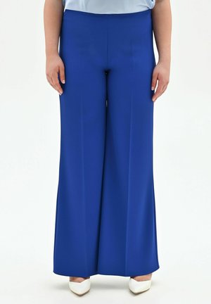 Femme portant un pantalon taille haute à jambes larges de couleur bleu roi, associé à des escarpins blancs à bouts pointus, debout devant un fond blanc.