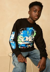 Musta pikkseelikuga T-särk, millel on värvilised Sonic the Hedgehogi graafilised kujutised siniste, roheliste ja valgete aktsentidega. Lahtine istuvus, puuvillane materjal.