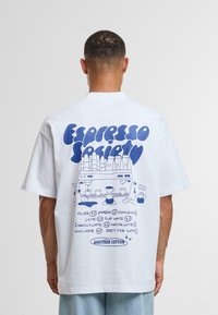 Camiseta de algodón blanca con un diseño gráfico azul en la parte posterior, que representa varias bebidas de café y una ilustración de una máquina de café.