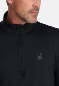 Zwarte pullover met een hoge kraag, voorzien van een halve rits en een subtiele spinnenlogo op de linkerkant van de borst. Gladde, rekbare stof.