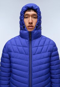 Modrá prošívaná péřová bunda s vysokou, nařasenou kapucí. Obsahuje horizontální a diagonální švy s tkaničkami u límce.