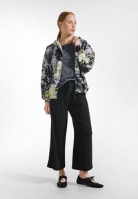 Lätt bomberjacka med blommönster i svart, vitt och gult över en grå skjorta; svarta byxor med vida ben; svarta flata skor med rosetter.