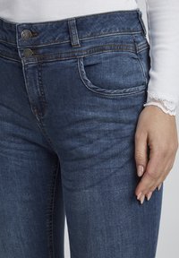 Tamsiai mėlyni džinsai su aukštu juosmeniu, dvigubu sagčiu, priekinėmis kišenėmis ir subtiliu įbrėžimo tekstūru šlaunų srityje.