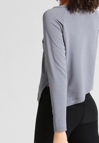 Maglia grigia a maniche lunghe con vestibilità ampia, caratterizzata da un orlo arrotondato, fessure laterali e un tessuto leggero e morbido. Abbinata a leggings neri.