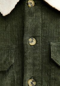 Camisa de pana verde oliva con un suave cuello crema, presenta cierres de botones y dos bolsillos en la parte frontal. Detalle de tela acanalada texturizada.