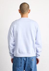 Light blue crewneck sweatshirt with long sleeves, ribbed cuffs, and hem. Soft texture, plain design, worn with blue denim jeans.