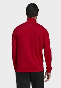 Red athletic jacket made of smooth, lightweight fabric. Features a high collar, long sleeves, and white accents along the shoulders.