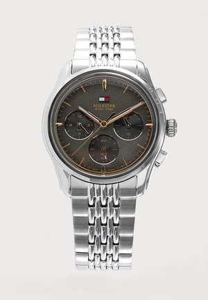 Montre-bracelet en métal argenté avec cadran noir affichant trois sous-cadrans, des marqueurs dorés et le logo "Hilfiger Dual Time" sous un drapeau rouge, blanc et bleu.
