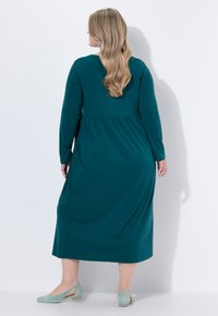 Teal mekko, jossa on pitkät hihat, gathered vyötärö, polven pituinen hame ja sileä tekstuuri. Yhdistetty vaaleanvihreisiin slingback-kenkiin.