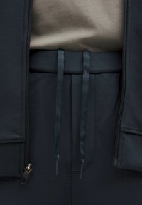 Mørkegrå jakke med lynlås foran, parret med højhævede mørke bukser med to stofsnore og en blød, tekstureret top.