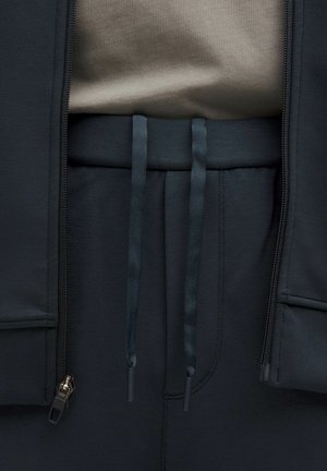 Ciemnoszara kurtka z zamkiem z przodu, w połączeniu z ciemnymi spodniami o wysokim stanie z dwoma sznurkami z materiału oraz miękką, teksturowaną bluzką.