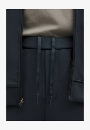 Ciemnoszara kurtka z zamkiem z przodu, w połączeniu z ciemnymi spodniami o wysokim stanie z dwoma sznurkami z materiału oraz miękką, teksturowaną bluzką.