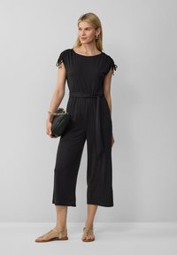 Schwarzer Jumpsuit mit kurzen Ärmeln, Kordelzug-Details, Taillengürtel, cropped weite Beine, kombiniert mit hellen Sandalen und einer kleinen Handtasche.