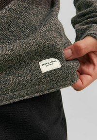 Grijze gebreide trui met een label waarop "JACK & JONES EST. 1990." staat. De stof is gestructureerd en zacht, met een ronde halslijn.