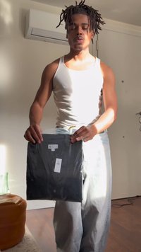 Jeune homme aux dreadlocks portant un débardeur blanc et un jean, tenant une chemise noire emballée à l'intérieur, avec la lumière du soleil sur lui.