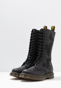 Černé kožené šněrovací boty s květinovou výšivkou, žlutým prošíváním a značkovými poutky, umístěné vedle sebe na bílé ploše.