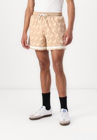 Béžové vzorované šortky s tahem v pase a bílým lemováním, doplněné bílými teniskami a černými ponožkami, zobrazené na stojícím modelu.
