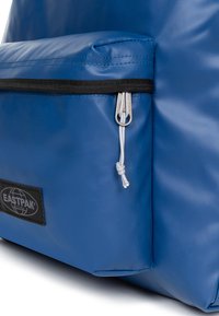 Blå Eastpak ryggsäck med ficka och svarta dragkedjesömmar, silverfärgad dragkedjehandtag och vit knut på dragkedjans flik.