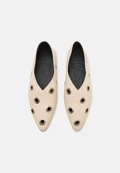 Beige leather loafers with a pointed toe, featuring large circular cutouts and metallic accents. Textured black insole with embossed branding.