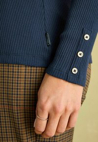 Námornícky pruhovaný top s dlhým rukávom a gombíkovými detailami na manžetách, skombinovaný s hnedými károvanými nohavicami. Texturovaná látka a strih prispôsobený postave.