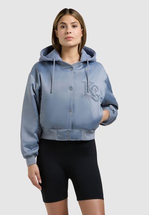 Kvinde iført en lyseblå hættetrøje med broderet logo og sorte cykelshorts, stående med den ene hånd i jakkenlommen.