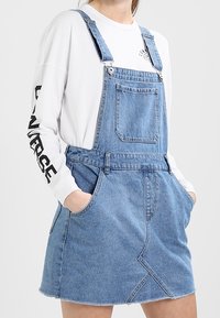 Denim overall klänning i ljusblått med justerbara remmar, frontficka och rå fåll. Har en triangulär sömdetalj på kjolen.