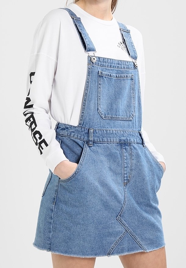 Denim overall klänning i ljusblått med justerbara remmar, frontficka och rå fåll. Har en triangulär sömdetalj på kjolen.