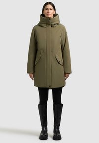 Olivgrüner langer Mantel mit hohem Kragen, zwei vorderen Taschen und Knopfverschluss. Getragen mit schwarzen Leggings und knielangen schwarzen Stiefeln.