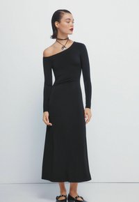 Zwart, langemouwen jurk met asymmetrische halslijn en aansluitende taille, die doorgaat naar een vloeiende zoom. Draag met een zwarte choker en platte schoenen.