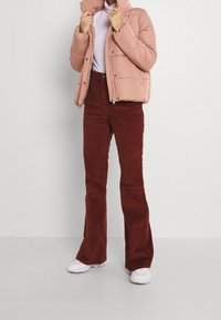 Roze gewatteerde jas, hoge kraag, ritssluiting, gecombineerd met roestkleurige corduroy flared broek. Witte sneakers maken de outfit af. Eenvoudige achtergrond.