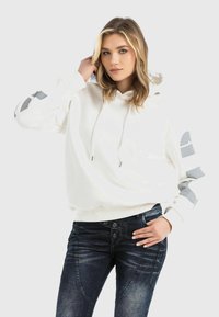 White hoodie with gray accents on sleeves, featuring a drawstring and ribbed hem. Paired with dark blue denim jeans.