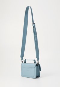 Lyseblå skinncrossbody-veske med justerbar stropp, liten topphåndtak og frontklaff. Har en subtil preget logo på forsiden.