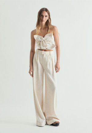 Mujer joven con cabello largo que lleva un top beige sin tirantes con un gran nudo y pantalones anchos de pierna alta en color crema y zapatos de plataforma.