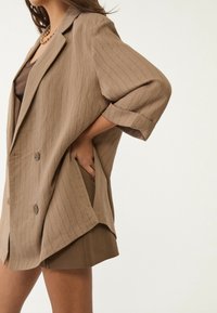 Vrouw in een lichtbruin gebroken streepjas met opgerolde mouwen, over een bruine top en bijpassende shorts, met een hand in de zak van de blazer.