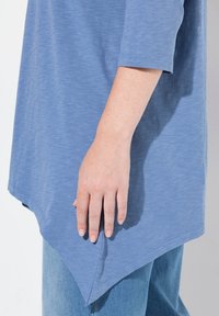 Vaaleansininen, pitkähihaisempi paita pyöreällä helman muotoilulla ja teksturoidulla kankaalla, yhdistettynä vaaleansinisiin farkkuihin, käsi hellästi paidalla.