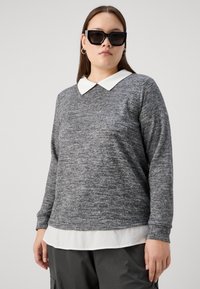 Suéter largo de manga larga texturizado en gris con un cuello blanco, superpuesto a una camisa blanca. Unas gafas de sol negras de gran tamaño completan el conjunto.