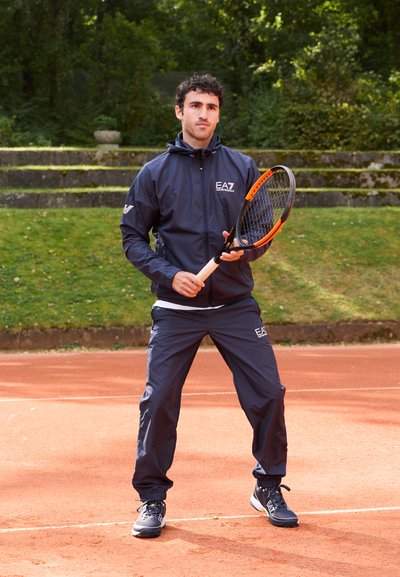 Морскосиньо tracksuit с цип и еластични панталони, съчетан с черни спортни обувки. Моделът държи тенис ракета с оранжев акцент.