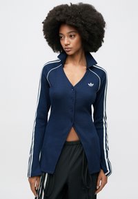 Tumši zila ribota sporta krekla ar garām piedurknēm un pogām, ar V veida kakla izgriezumu, baltām sānu strīpām un izšūtu logotipu uz kreisās krūts puses.