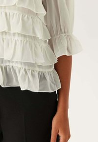 Blusa bianca con volant in tessuto trasparente sovrapposto, maniche a palloncino e polsini elasticizzati, abbinata a pantaloni neri. Primo piano della manica e della mano.
