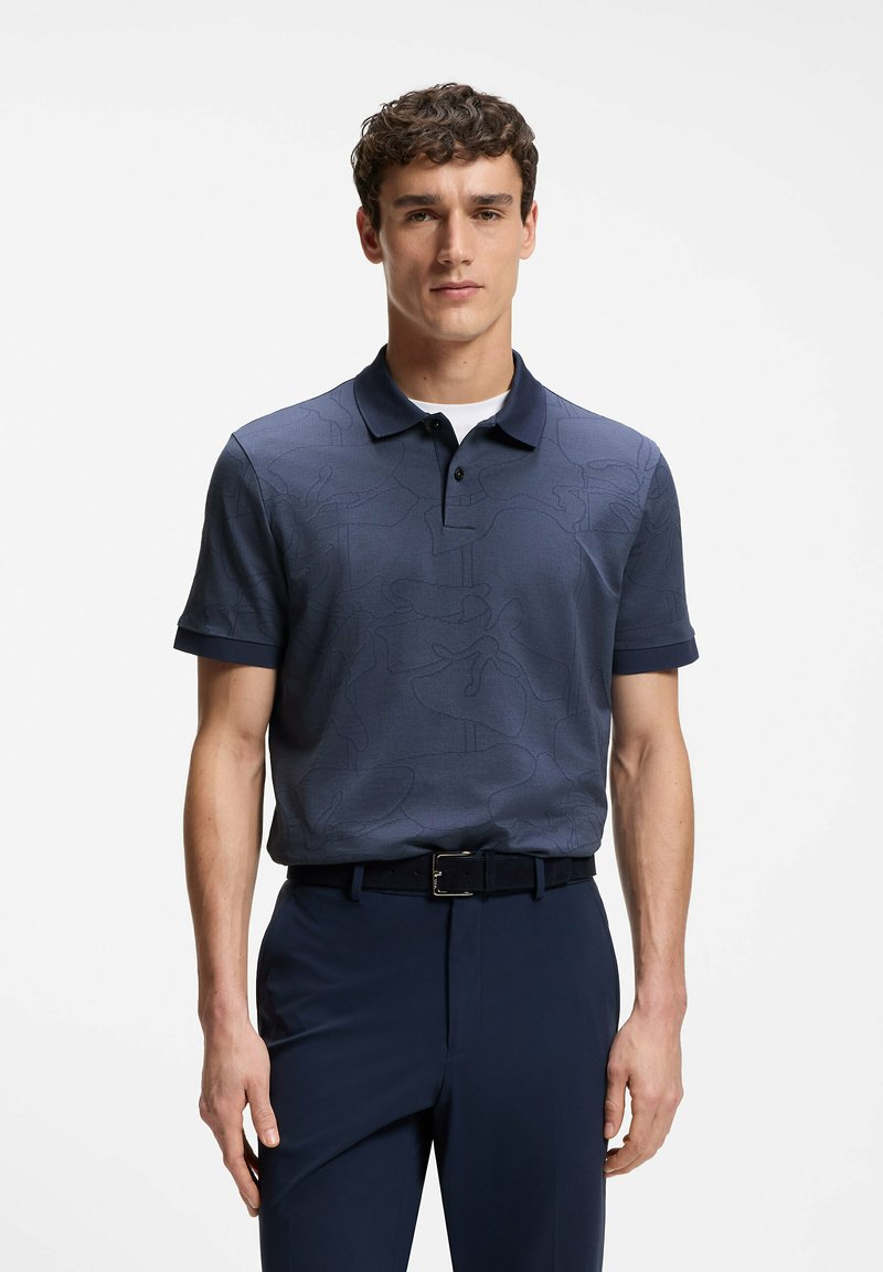 Homme aux cheveux courts et bouclés, portant un polo bleu marine à motifs et un pantalon bleu marine assorti avec une ceinture noire, debout devant un fond blanc.