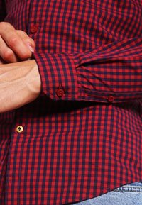 Persoon die de manchetknop vastmaakt van een rood en zwart geblokt overhemd met lange mouwen, in een spijkerbroek.