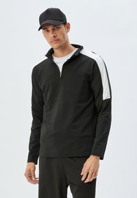 Top atlético negro con cremallera y acentos blancos en los hombros. Cuenta con mangas largas y un cuello alto, hecho de una tela suave y elástica.