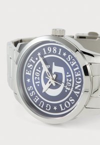 Reloj de acero inoxidable plateado con un dial de color azul oscuro, que presenta texto blanco, manecillas metálicas y acentos de logo. Pulsera ajustable de eslabones.
