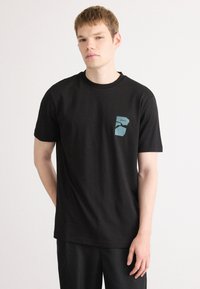 Schwarzes T-Shirt aus Baumwolle mit Rundhalsausschnitt, mit einem blauen Grafiklogo auf der linken Brustseite. Kurze Ärmel, lockere Passform.
