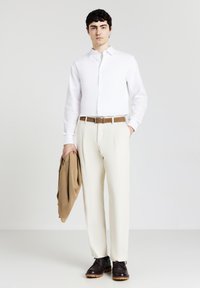 Camicia bianca con bottoni, pantaloni color crema, cintura marrone, maglione marrone appoggiato sul braccio e scarpe marroni scure con suola resistente. Design minimalista.