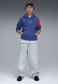 Felpa blu con accenti rossi e blu, tasca a marsupio, abbinata a jeans baggy chiari, dettaglio in catena argentata, calzature nere e berretto nero.